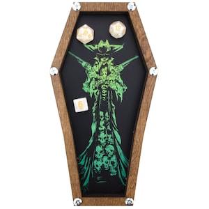 May include: A wooden coffin-shaped dice tray with a green and black illustration of a skeletal figure holding two pistols. The tray has a black felt interior and includes four dice with gold numbers.