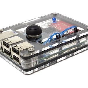 May include: A clear acrylic case houses a small electronic device with a black joystick, ports, and a red circuit board. Blue and black wires are visible inside the case. The device is secured with silver screws and black rubber bands.