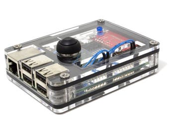 May include: A transparent acrylic case containing a small electronic device with a black joystick, ports, and a red circuit board. Blue and black wires are visible. The device is secured with silver screws and black rubber bands.