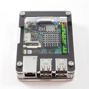May include: A black and clear plastic case with a circuit board inside. The board has a green and black color scheme and includes a heatsink, a network port, and a row of pins. The case has the text "H6388" and "H6388" printed on it.