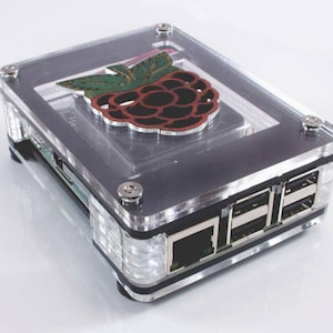 May include: A clear acrylic case for a Raspberry Pi computer. The case has a black base and a clear top with a raspberry logo on it. The case has a USB port and an Ethernet port on the side.
