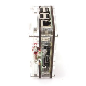 Op de afbeelding: Een transparante acrylbehuizing met een houten paneel aan de zijkant. De behuizing heeft verschillende poorten, waaronder een HDMI-poort, een Ethernet-poort en audio-aansluitingen.
