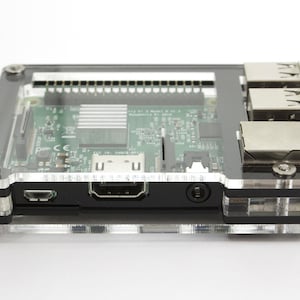 Peut inclure: Un boîtier transparent en acrylique pour un ordinateur Raspberry Pi. Le boîtier a une base noire et un dessus transparent, permettant de voir le circuit imprimé vert du Raspberry Pi. Le boîtier possède un port USB, un port HDMI et un port Ethernet.