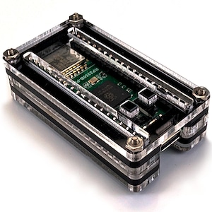 May include: A rectangular electronic development board encased in a layered black and clear acrylic housing. The device features a green circuit board with a central chip labelled RP2350B-P, two black push buttons, and a USB-C port, secured by metal screws.