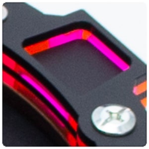 Op de afbeelding: Close-up van een zwart metalen object met een roze en oranje LED-lampje. Het lampje heeft de vorm van een vierkant en is omgeven door een zwarte rand.