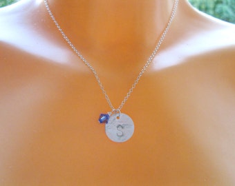 May include: A silver necklace with a small round pendant featuring the letter "S". A small blue crystal dangles from the pendant.