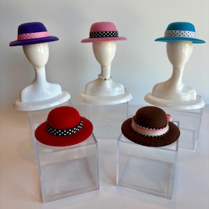 May include: Five colorful hats on display. The hats are displayed on white mannequin heads. The hats are in various colors, including purple, pink, red, brown, and teal. Each hat has a contrasting band with polka dots or stripes.