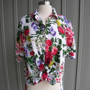 May include: White button-down shirt with a vibrant floral pattern featuring red, pink, purple, and yellow flowers. The short-sleeved shirt is knotted at the waist, showcasing a casual style. The shirt is displayed on a mannequin.