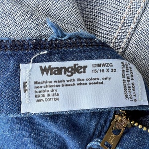 May include: Close-up of blue denim jeans with a visible zipper and a white tag. The tag reads "Wrangler" and includes washing instructions and material information. The jeans are made of 100% cotton.