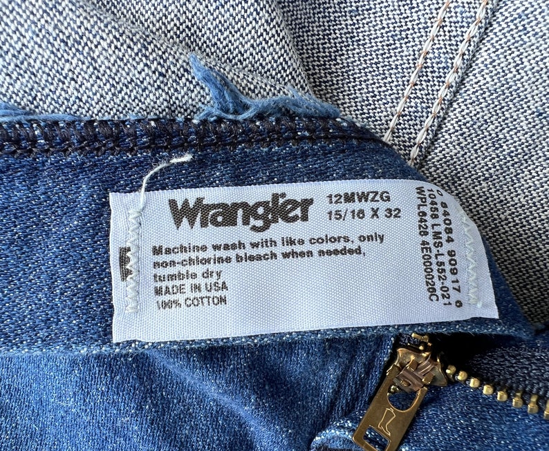 May include: Close-up of blue denim jeans with a visible zipper and a white tag. The tag reads "Wrangler" and includes washing instructions and material information. The jeans are made of 100% cotton.