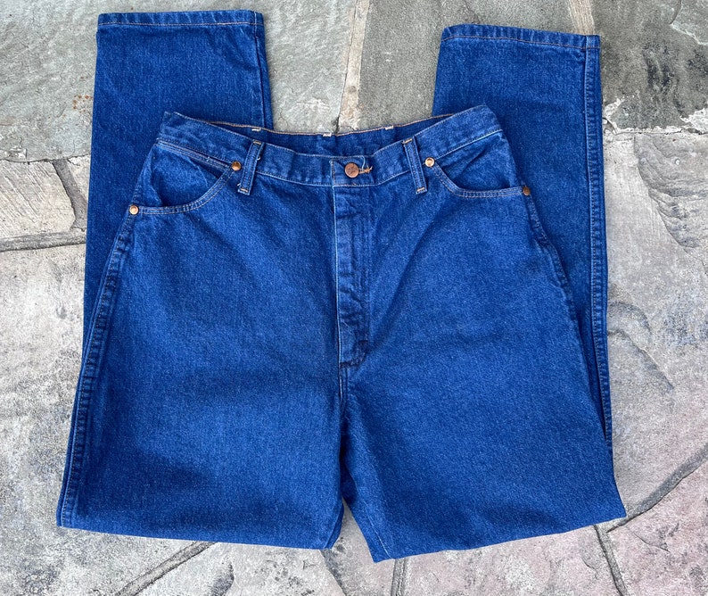 May include: A pair of classic, high-waisted, dark blue denim jeans. The jeans feature a button and zipper closure, front and back pockets, and copper-colored rivets. The straight-leg design is visible in this close-up shot.
