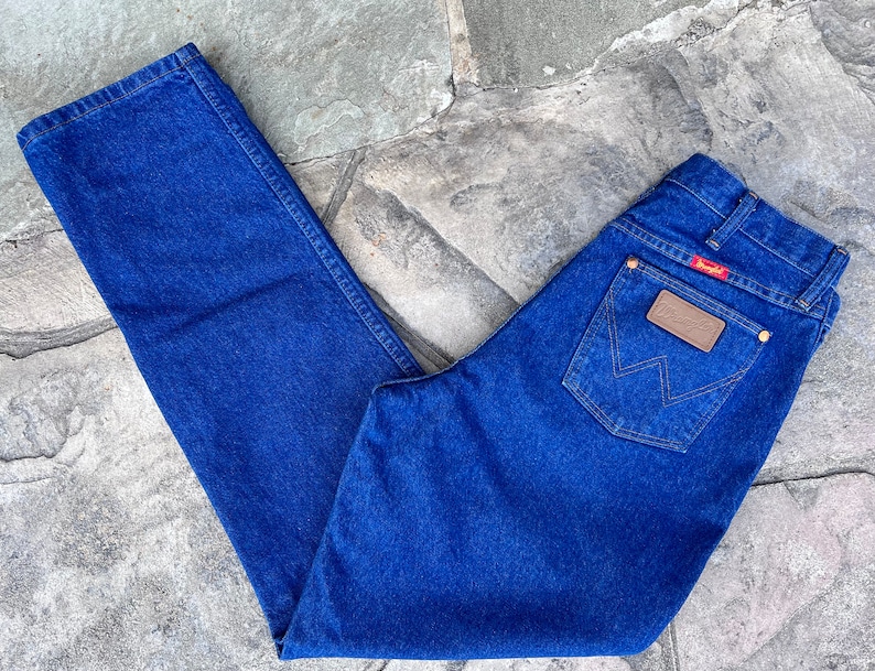 May include: Classic blue denim jeans with a straight leg cut. The jeans feature a brown leather patch with the Wrangler logo on the back pocket and a red tag. The jeans are laid out on a stone surface.