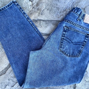 May include: Blue denim jeans with a classic five-pocket design. The jeans have a faded wash and a straight leg cut. The back pocket features a decorative stitch. The jeans are folded on a stone surface.