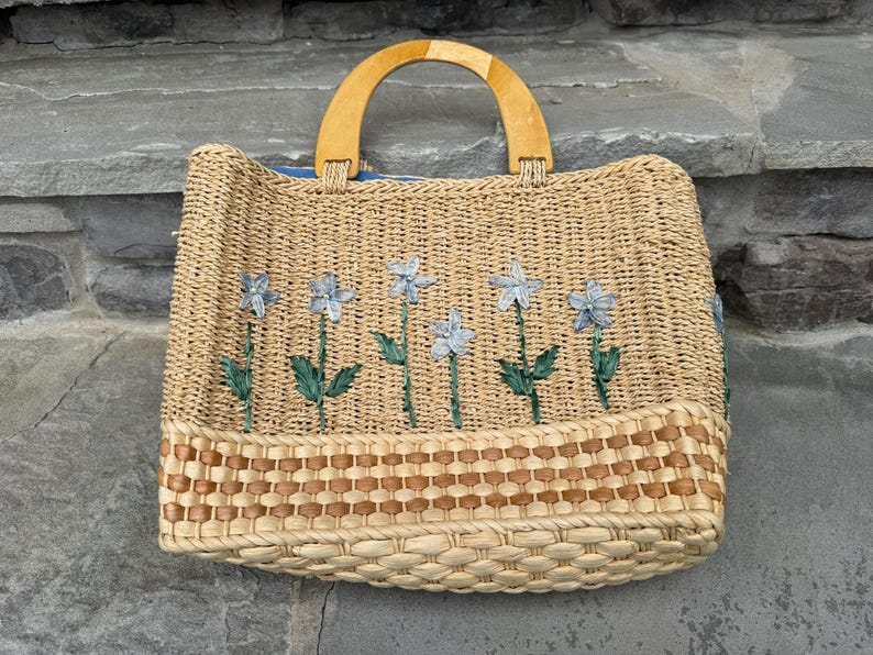 K&ouml;nnte beinhalten: Gewebte Stroh-Tragetasche mit Holzgriff. Die Tasche hat gestickte blaue Blumen mit gr&uuml;nen Stielen und Bl&auml;ttern. Der Boden der Tasche hat ein gewebtes braunes und beiges Muster.