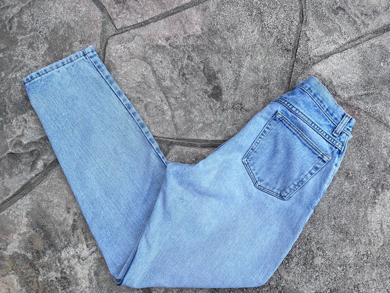May include: Light blue denim jeans with a classic five-pocket design. The jeans are folded and laid on a textured gray surface. The jeans have a straight leg cut and a button and zipper closure.