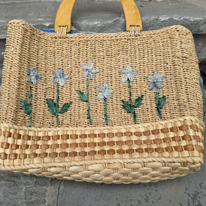 K&ouml;nnte beinhalten: Gewebte Stroh-Tragetasche mit Holzgriffen. Die Tasche hat ein Blumendesign mit blauen Blumen und gr&uuml;nen Stielen. Der Boden der Tasche hat ein gewebtes braunes Muster. Die Tasche ist zum Tragen von Gegenst&auml;nden konzipiert.