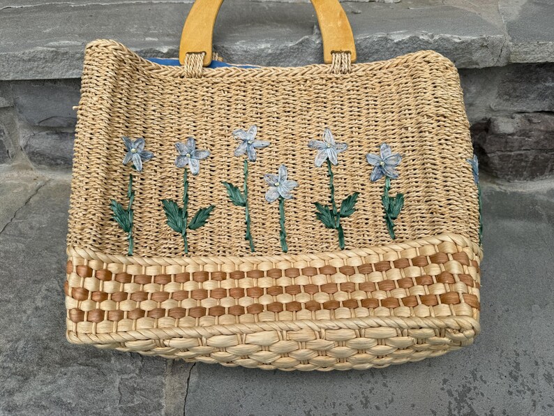 K&ouml;nnte beinhalten: Gewebte Stroh-Tragetasche mit Holzgriffen. Die Tasche hat ein Blumendesign mit blauen Blumen und gr&uuml;nen Stielen. Der Boden der Tasche hat ein gewebtes braunes Muster. Die Tasche ist zum Tragen von Gegenst&auml;nden konzipiert.