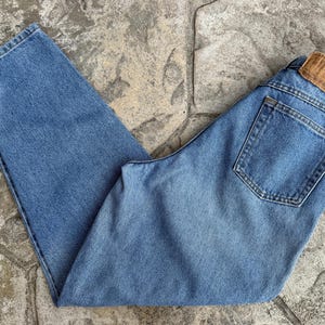 May include: Light blue denim jeans with a classic five-pocket design. The jeans have a straight leg cut and a leather patch on the back waistband. The jeans are folded on a stone surface.