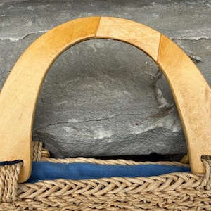 K&ouml;nnte beinhalten: Nahaufnahme einer gewebten Strohtasche mit einem hellbraunen Holzgriff. Der Griff ist gew&ouml;lbt und mit geflochtenem Seil an der Tasche befestigt. Ein blauer Stoff s&auml;umt das Innere der Tasche.