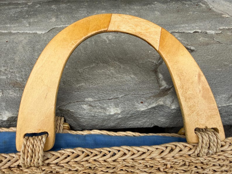 K&ouml;nnte beinhalten: Nahaufnahme einer gewebten Strohtasche mit einem hellbraunen Holzgriff. Der Griff ist gew&ouml;lbt und mit geflochtenem Seil an der Tasche befestigt. Ein blauer Stoff s&auml;umt das Innere der Tasche.