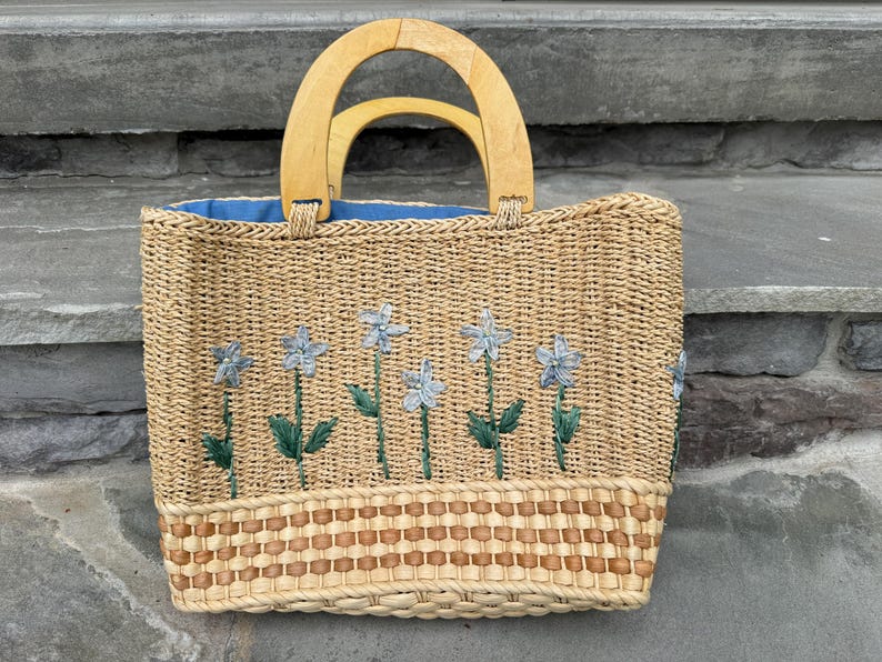 K&ouml;nnte beinhalten: Gewebte Stroh-Tragetasche mit hellblauem Innenraum und Holzgriffen. Die Tasche hat gestickte hellblaue Blumen mit gr&uuml;nen Stielen und Bl&auml;ttern. Der Boden der Tasche hat ein gewebtes braunes und beiges Muster.