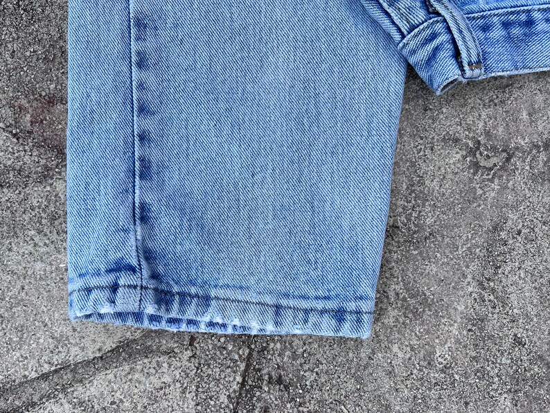 May include: Close-up of light blue denim jeans. The image shows the hem of the jeans, the stitching, and the texture of the fabric. The jeans are slightly faded, suggesting wear.