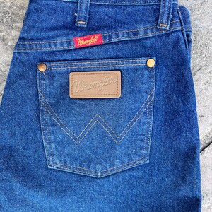 May include: Close-up of blue denim jeans with a back pocket featuring a brown leather patch with the brand name "Wrangler". The jeans have a red tag with the "Wrangler" logo and brass-colored rivets. The stitching is a contrasting tan color.