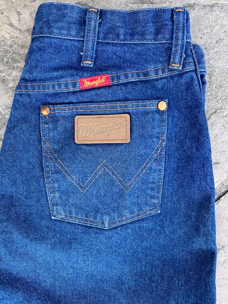 May include: Close-up of blue denim jeans with a back pocket featuring a brown leather patch with the brand name "Wrangler". The jeans have a red tag with the "Wrangler" logo and brass-colored rivets. The stitching is a contrasting tan color.