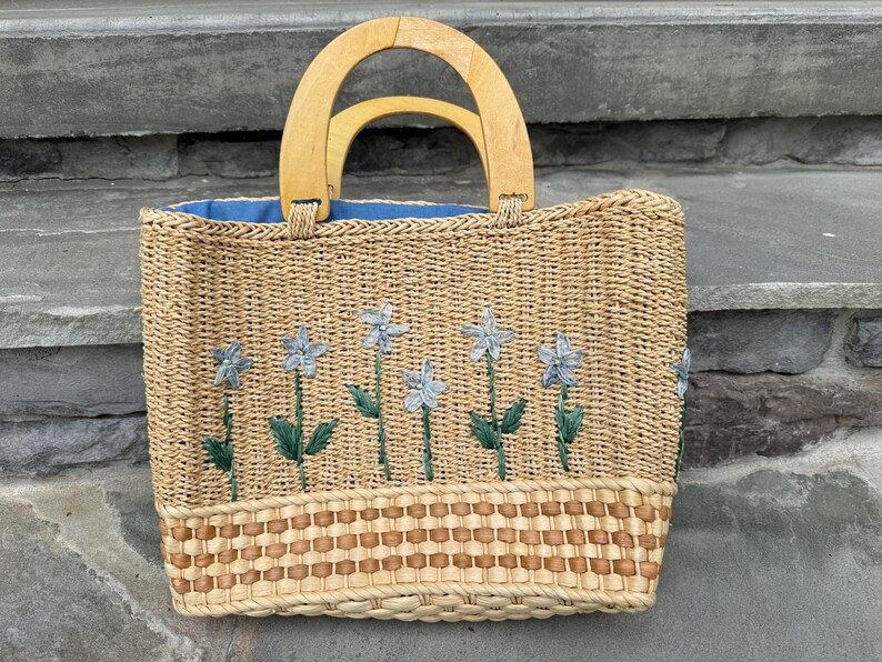 K&ouml;nnte beinhalten: Gewebte Stroh-Tragetasche mit Holzgriffen. Die Tasche hat gestickte blaue Blumen mit gr&uuml;nen Stielen und Bl&auml;ttern. Der Innenraum ist mit blauem Stoff gef&uuml;ttert. Der Boden der Tasche hat ein dekoratives Webmuster.