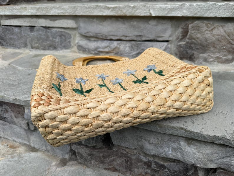 K&ouml;nnte beinhalten: Gewebte Stroh-Tragetasche mit rechteckiger Form und Holzgriff. Die Tasche hat gestickte blaue Blumen mit gr&uuml;nen Stielen und Bl&auml;ttern. Die Tasche ist aus naturfarbenem Stroh gefertigt.