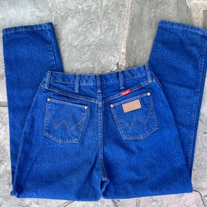May include: Blue denim jeans with a classic five-pocket design. The jeans feature the Wrangler logo on a leather patch and a red tag. The back pockets have a signature W-shaped stitching. The jeans are made of a durable denim fabric.