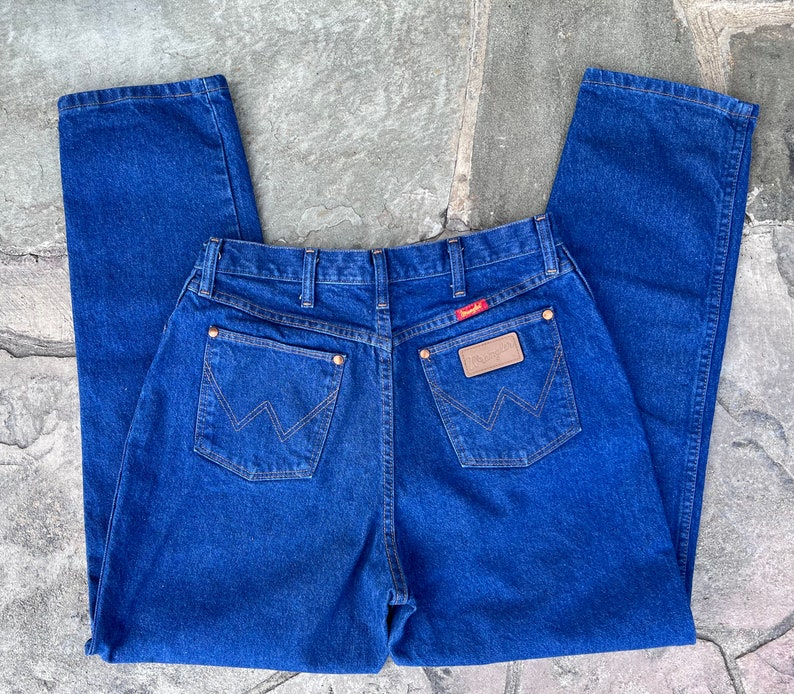May include: Blue denim jeans with a classic five-pocket design. The jeans feature the Wrangler logo on a leather patch and a red tag. The back pockets have a signature W-shaped stitching. The jeans are made of a durable denim fabric.