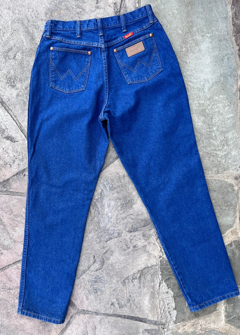 May include: Blue denim jeans with the Wrangler logo on the back pocket. The jeans have copper-colored rivets and a leather patch. The back pockets feature the Wrangler 'W' stitching.