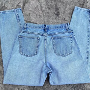 May include: Light blue denim jeans with a classic five-pocket design. The jeans are laid flat, showcasing the back pockets and the overall light wash. The fabric appears to be a medium weight denim.