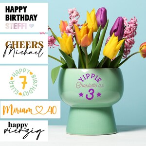 Könnte beinhalten: Ein grüner Keramiktopf mit weißem Sockel, verziert mit dem Text "YIPPIE Charlotte ist *3*" in Lila und Rosa. Der Topf ist mit einem Strauß gelber und lila Tulpen und rosa Hyazinthen gefüllt.