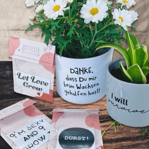 May include: Two white ceramic flower pots with black lettering. The first pot has a daisy plant and says "DANKE, dass Du mir beim Wachsen geholfen hast!" The second pot has a green plant and says "I will survive".