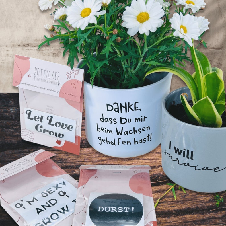 May include: Two white ceramic flower pots with black lettering. The first pot has a daisy plant and says "DANKE, dass Du mir beim Wachsen geholfen hast!" The second pot has a green plant and says "I will survive".