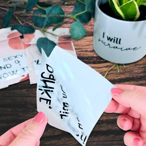 May include: A white vinyl sticker with the text "You will survive.  Oh, like?" in black lettering. The sticker is being peeled off of a clear transfer sheet.