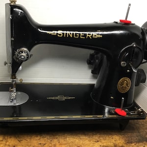 May include: A vintage black Singer sewing machine featuring gold decorative scrollwork along the base and the Singer logo in gold lettering on the arm. The machine includes a silver needle plate, a hand wheel, and two small red circular accents.