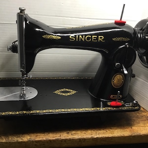 May include: A vintage black Singer sewing machine featuring gold filigree scrollwork along the base and arm. The machine includes a chrome needle bar, a large handwheel on the right side, and a circular gold emblem on the main body.