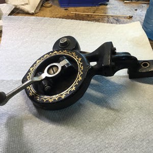 May include: A vintage black metal sewing machine hand crank attachment featuring a circular housing with decorative gold filigree patterns, a silver-toned metal handle with a dark wooden knob, and a mounting arm with two circular screw holes.