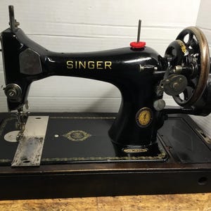 May include: A vintage black Singer sewing machine with gold decorative scrollwork, a hand crank on the side, and a metal needle plate. The machine features a prominent gold Singer logo on the arm and a circular emblem on the base.