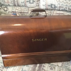 May include: A rectangular, dark brown wooden sewing machine carrying case with a smooth, polished finish. The case features a curved top, a metal handle attached to the centre, and the word SINGER printed in gold lettering on the front panel.