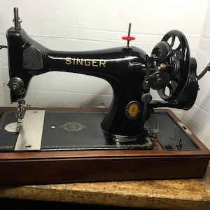 Peut inclure: Une machine à coudre Singer noire vintage avec des lettres dorées et des motifs décoratifs, montée sur une base en bois rectangulaire. La machine dispose d'une manivelle sur le côté droit, d'une plaque à aiguille argentée et d'un embout rouge.