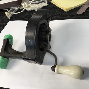May include: A vintage, black metal hand-cranked mechanism with a white handle. The mechanism has a complex design with visible gears and a crank arm. Green tape is applied to the base. The object is likely a part of a larger machine.