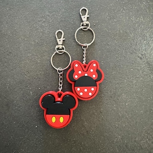 Może przedstawiać: Dwa breloczki do kluczy z motywami Myszki Miki i Myszki Minnie. Brelok Myszki Miki ma czerwony kontur, czarną głowę i żółte owalne oczy. Brelok Myszki Minnie ma czerwoną kokardę w białe grochy. Oba mają srebrne kółka.