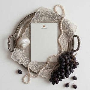 Pode incluir: Um cartão de papelaria retangular branco com um pequeno ícone de concha e o texto The Hansons, colocado numa bandeja de prata com um naperon de renda branca, um colar de pérolas, uma grande concha iridescente e uvas roxas escuras.