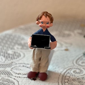 May include: A handcrafted figurine of a person holding a black laptop. The figure is wearing a navy blue shirt, khaki pants, and burgundy shoes. The figurine has brown hair and is set against a neutral background.