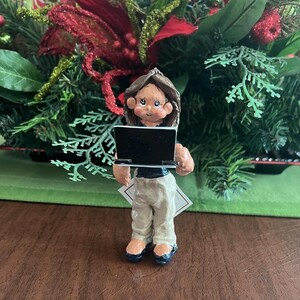 May include: A figurine of a person holding a black laptop, wearing a black top and tan pants. The figurine has brown hair and blue eyes. The background features a Christmas floral arrangement with red and green accents.