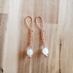 Puede incluir: Un par de pendientes de oro rosa con un colgante de piedra blanca en cada pendiente. Los pendientes cuelgan de una cadena delicada.
